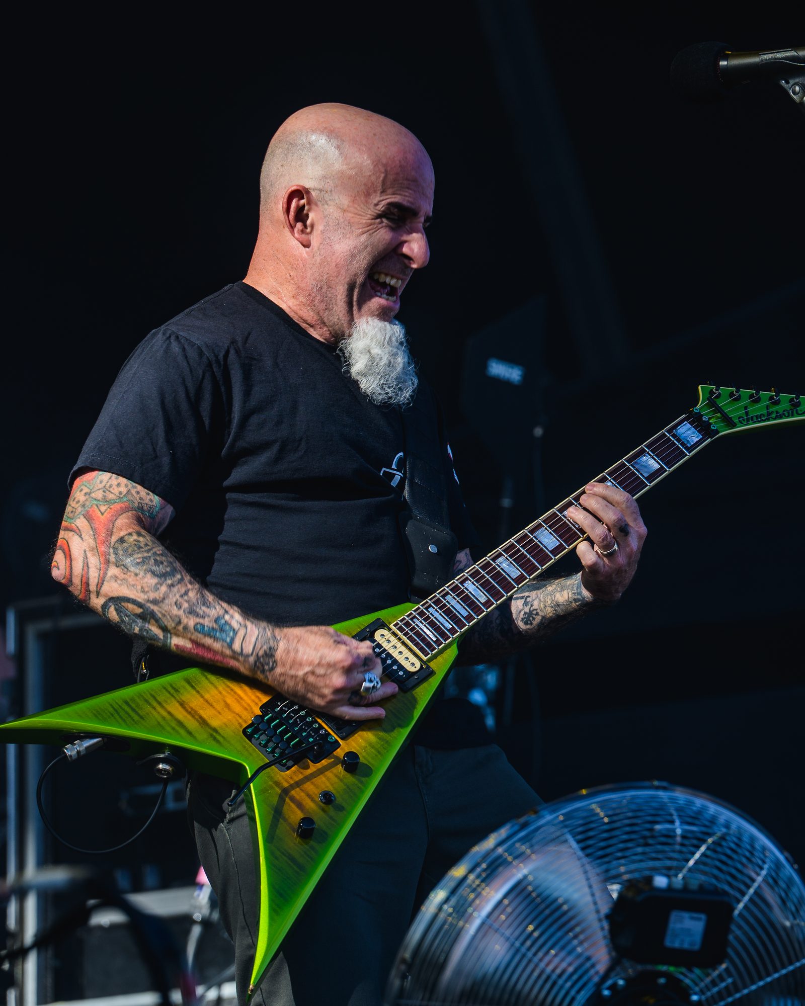 Scott Ian, bald with a gray goatee and tattooed arms, laughing as he plays a green-and-orange Jackson V guitar on an outdoor stage