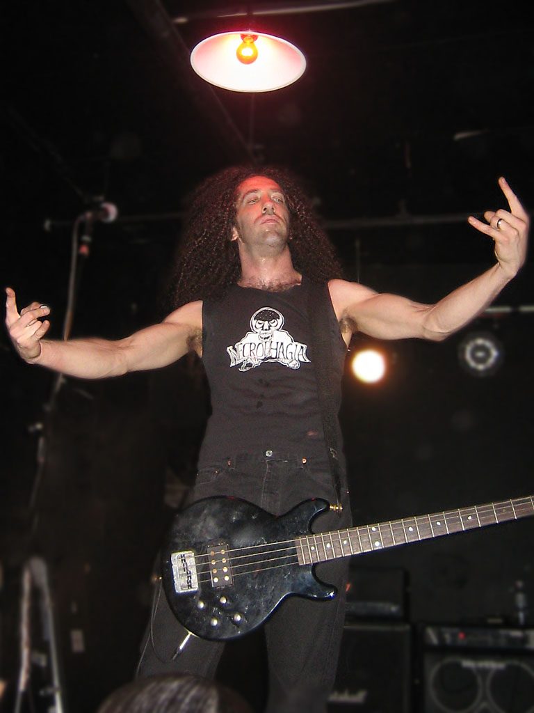 Dan Lilker on a small club stage under a hanging lamp, arms spread wide, long curly hair, a black bass guitar slung low and a Necrophagia shirt