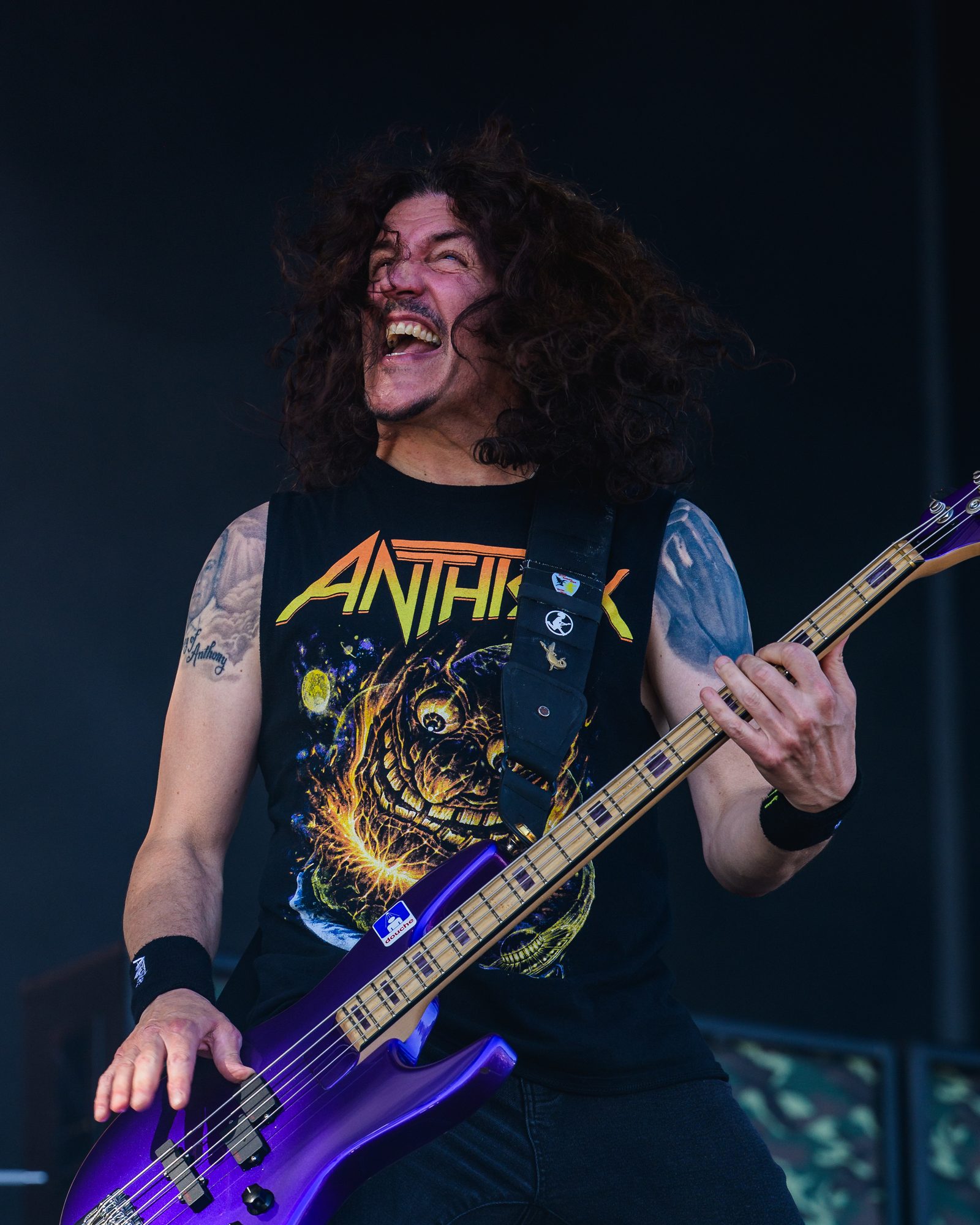 Frank Bello laughing with his head thrown back and curly hair flying, playing a purple bass, wearing a sleeveless Anthrax shirt with a cosmic monster print