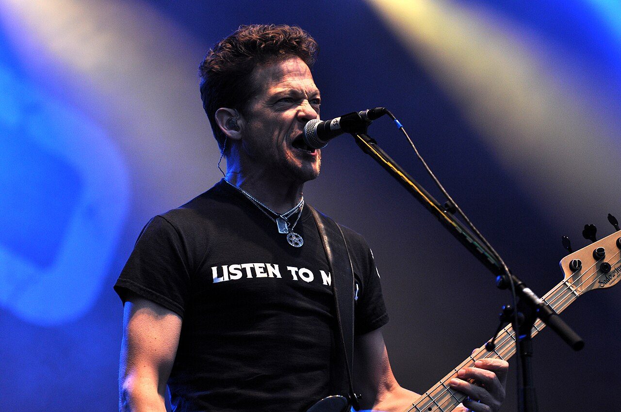 Jason Newsted singing into a microphone and playing a bass guitar on stage, wearing a black T-shirt reading Listen To Me, with blue and yellow stage lights behind him