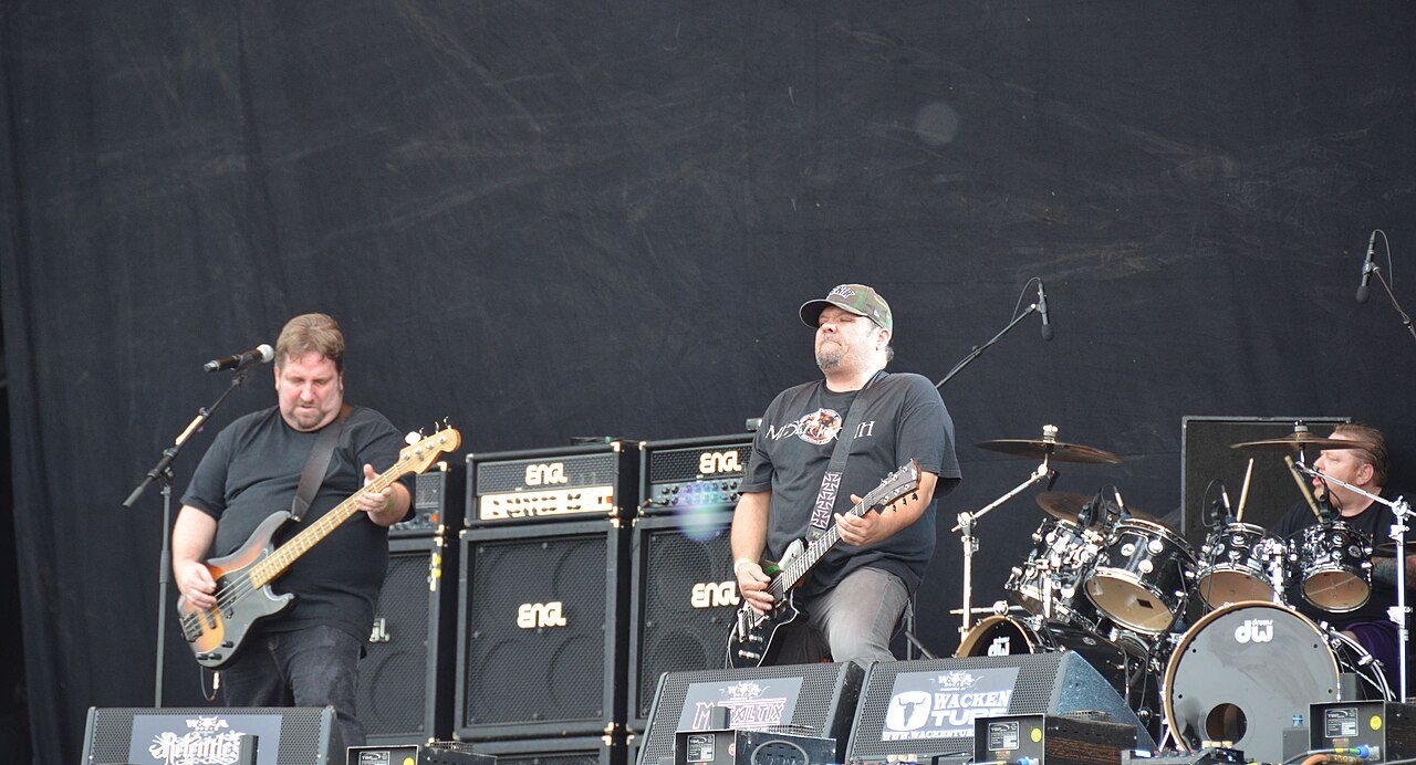 Sacred Reich on a large daytime festival stage, a bassist in black at the left mic, a guitarist in a cap beside him and a drummer behind his kit at the right, with a row of amplifier stacks and a black backdrop