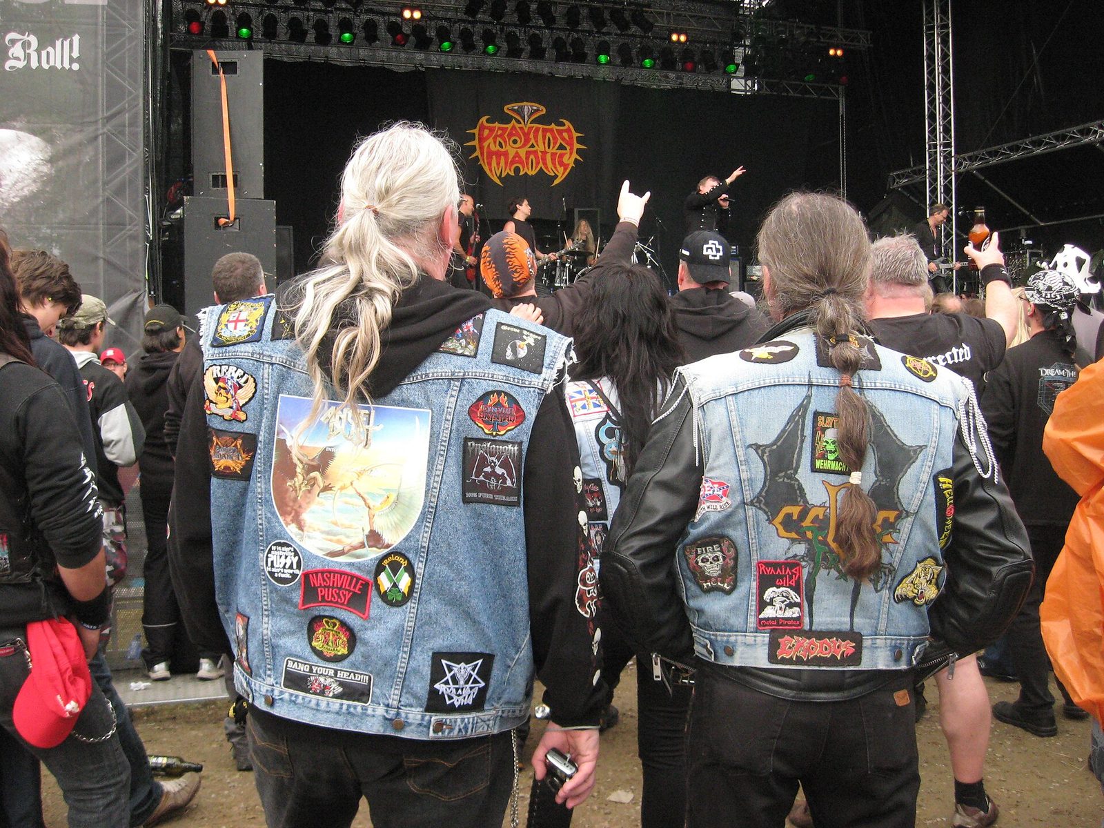 Two long-haired metal fans seen from behind at an outdoor festival, both in sleeveless blue denim vests covered in band patches, including Exodus, Slayer, Onslaught and Running Wild, watching Praying Mantis on stage