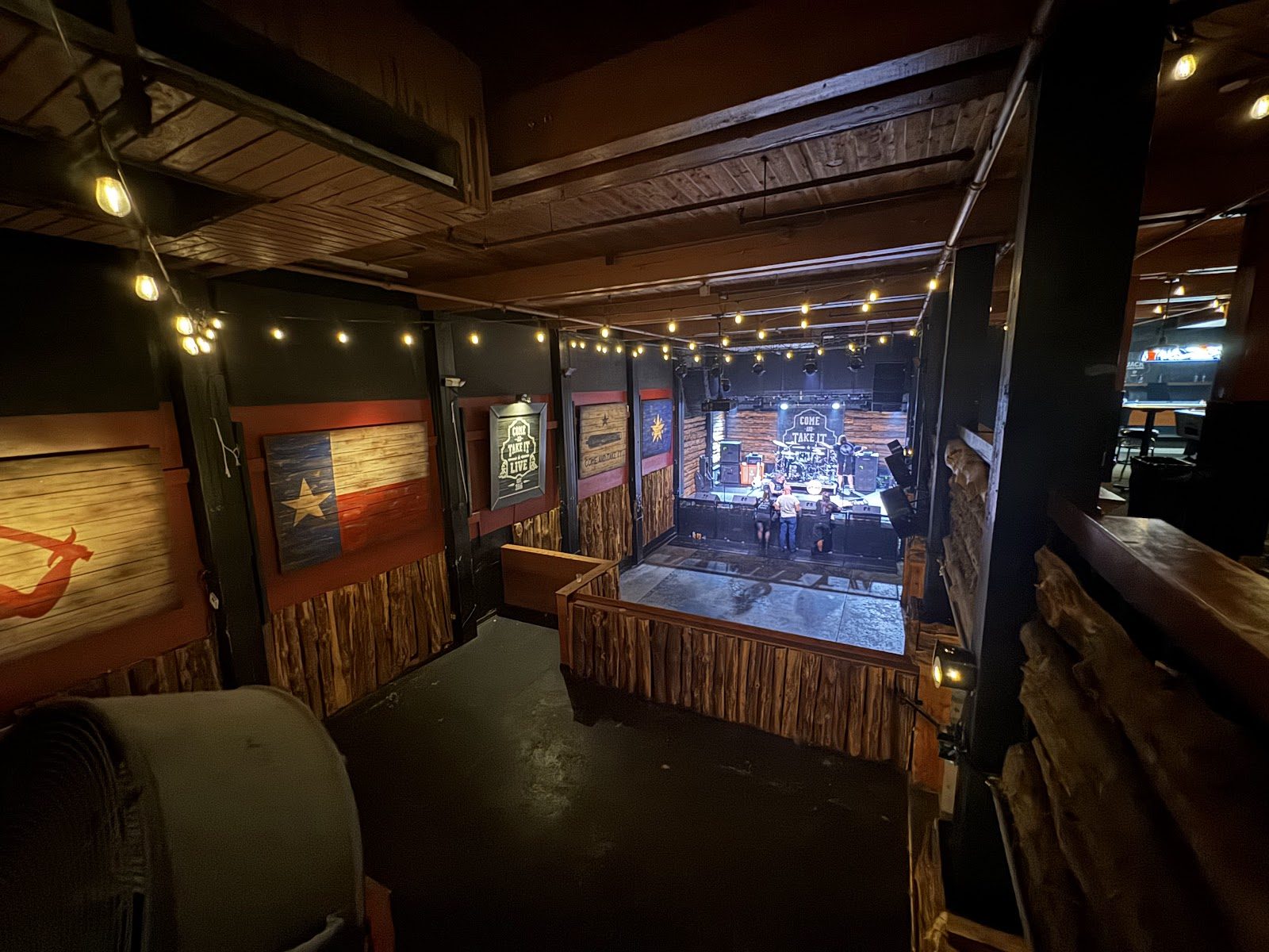 View from the upper level of Come and Take It Live down to the stage, wood-paneled walls with a Texas flag, string lights along the ceiling