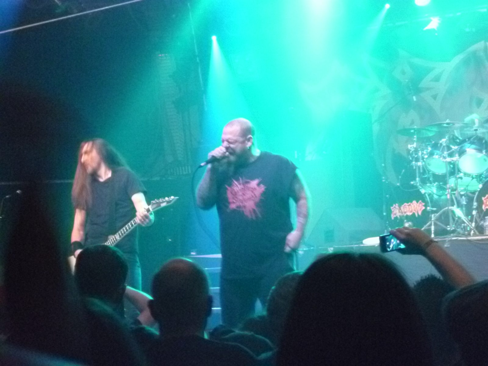 Exodus onstage under green light, a bald singer in a black T-shirt at the microphone beside a long-haired guitarist, the drum kit behind them