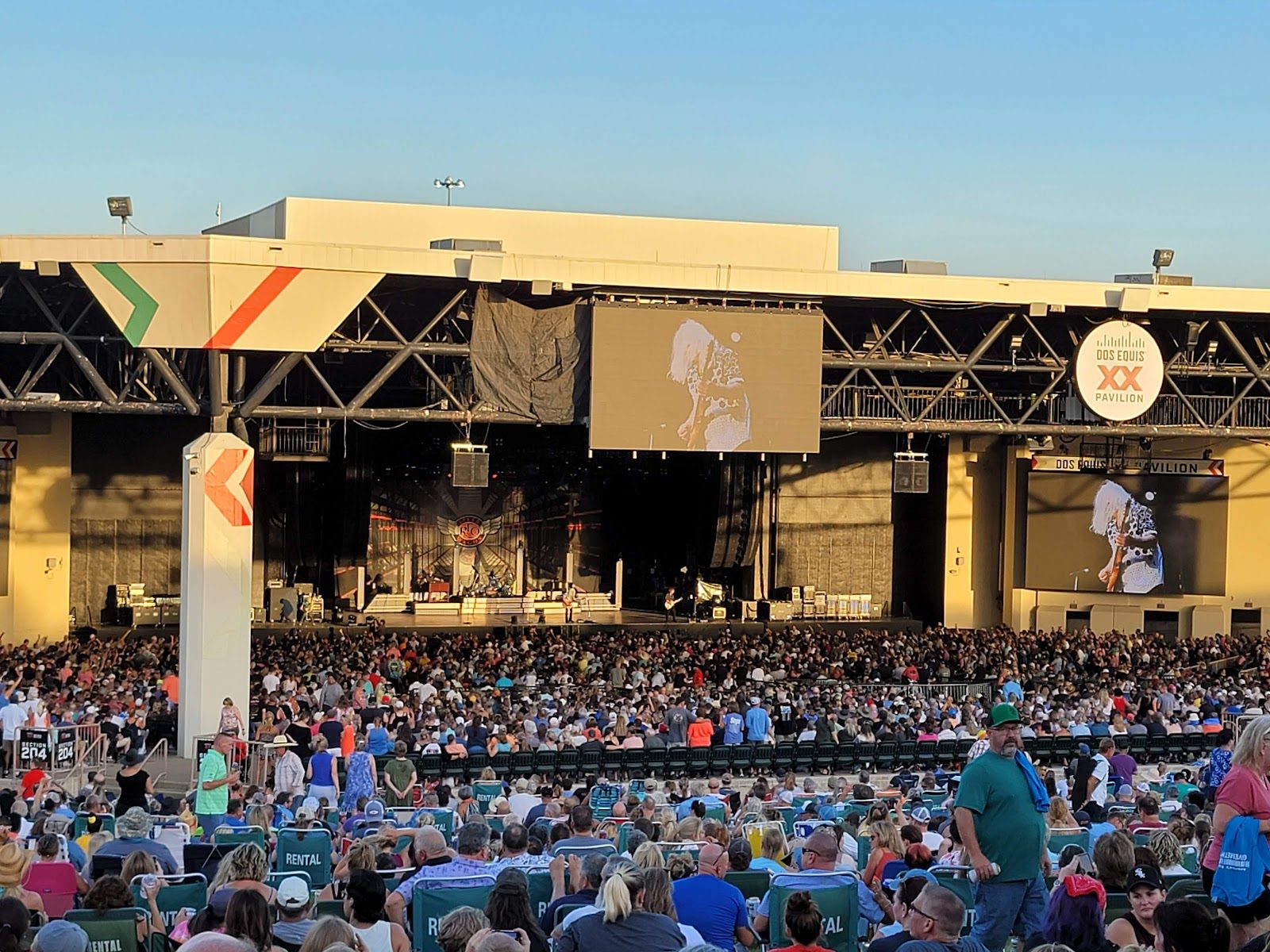 A packed outdoor amphitheater at golden hour, the crowd filling the seats and lawn in front of a covered stage with video screens on each side and a Dos Equis logo on the roof truss