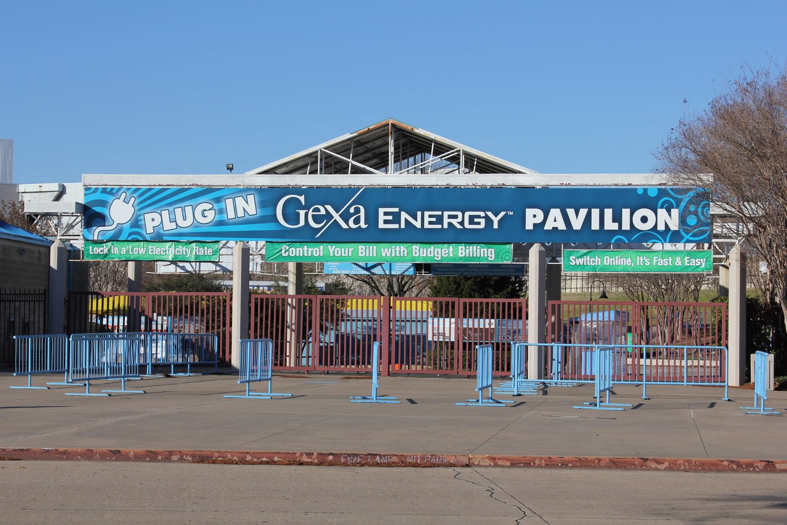 The east entrance gate of the Dallas amphitheater under a blue banner reading Gexa Energy Pavilion, with empty crowd barriers in front