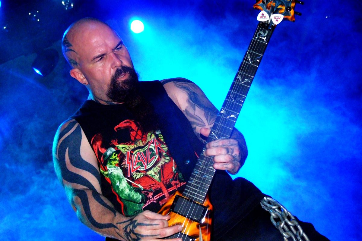 Kerry King, bald with a long dark beard and tattooed arms, playing a flame-finished guitar in a sleeveless Slayer shirt under blue stage light
