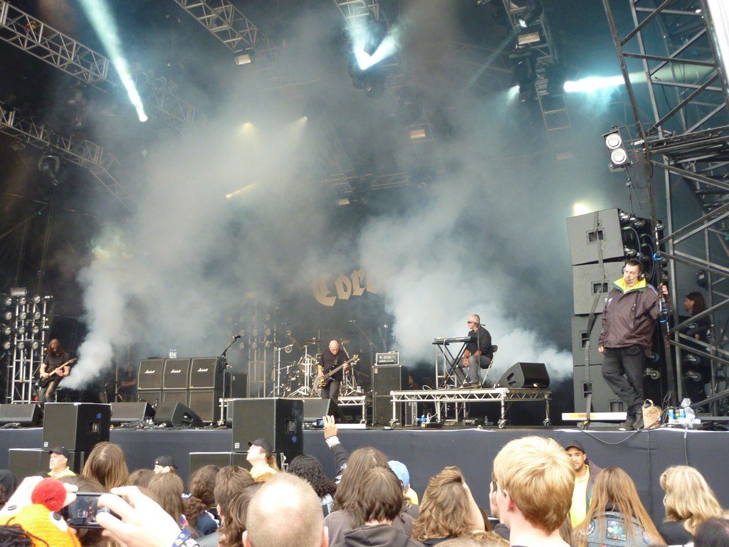 Coroner on a large outdoor festival stage wrapped in white smoke, the band's logo on the backdrop and a crowd of fans in the foreground