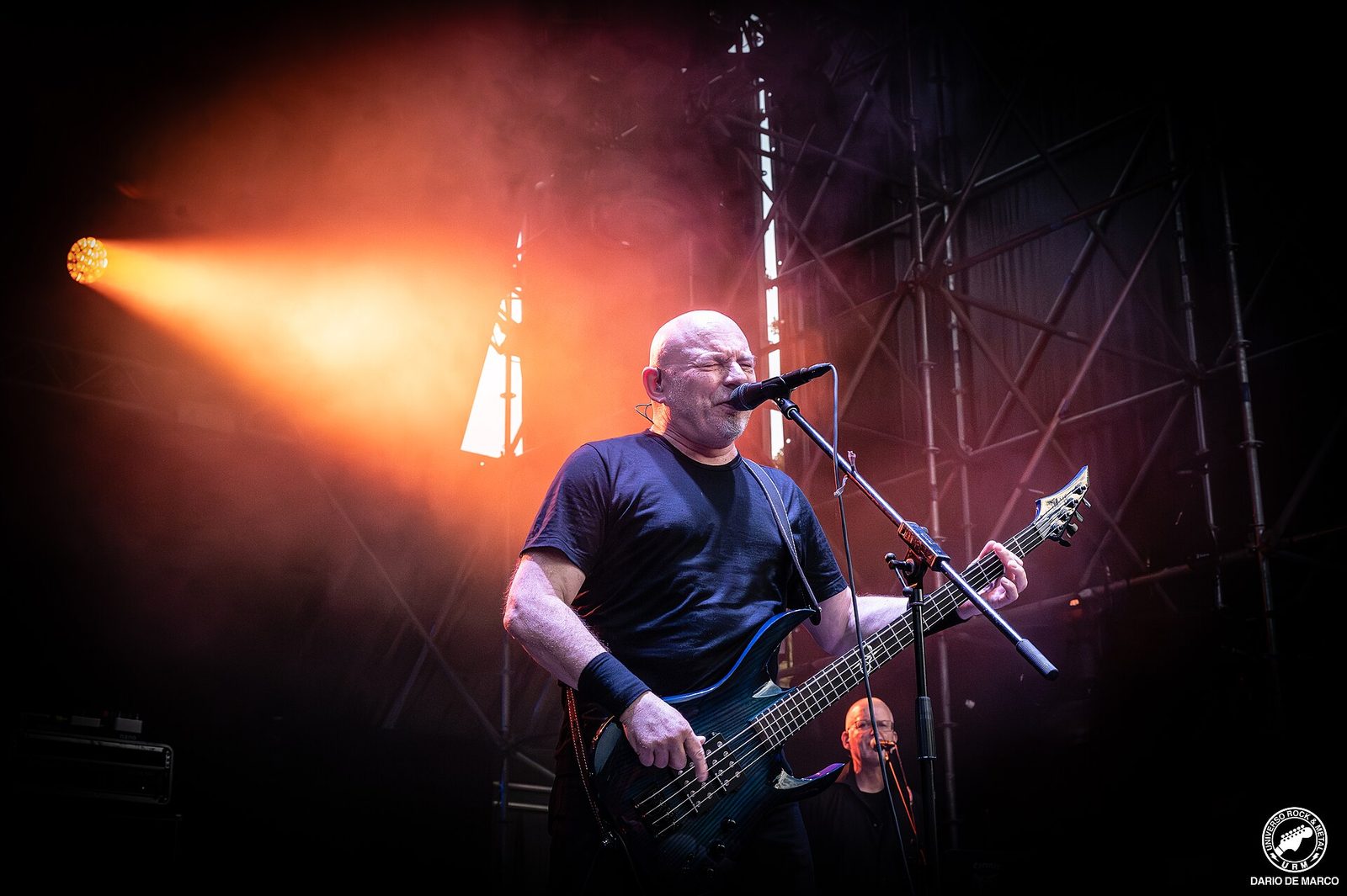A bald bassist in a black T-shirt singing into a microphone while playing a black bass, lit by an orange spotlight on a festival stage