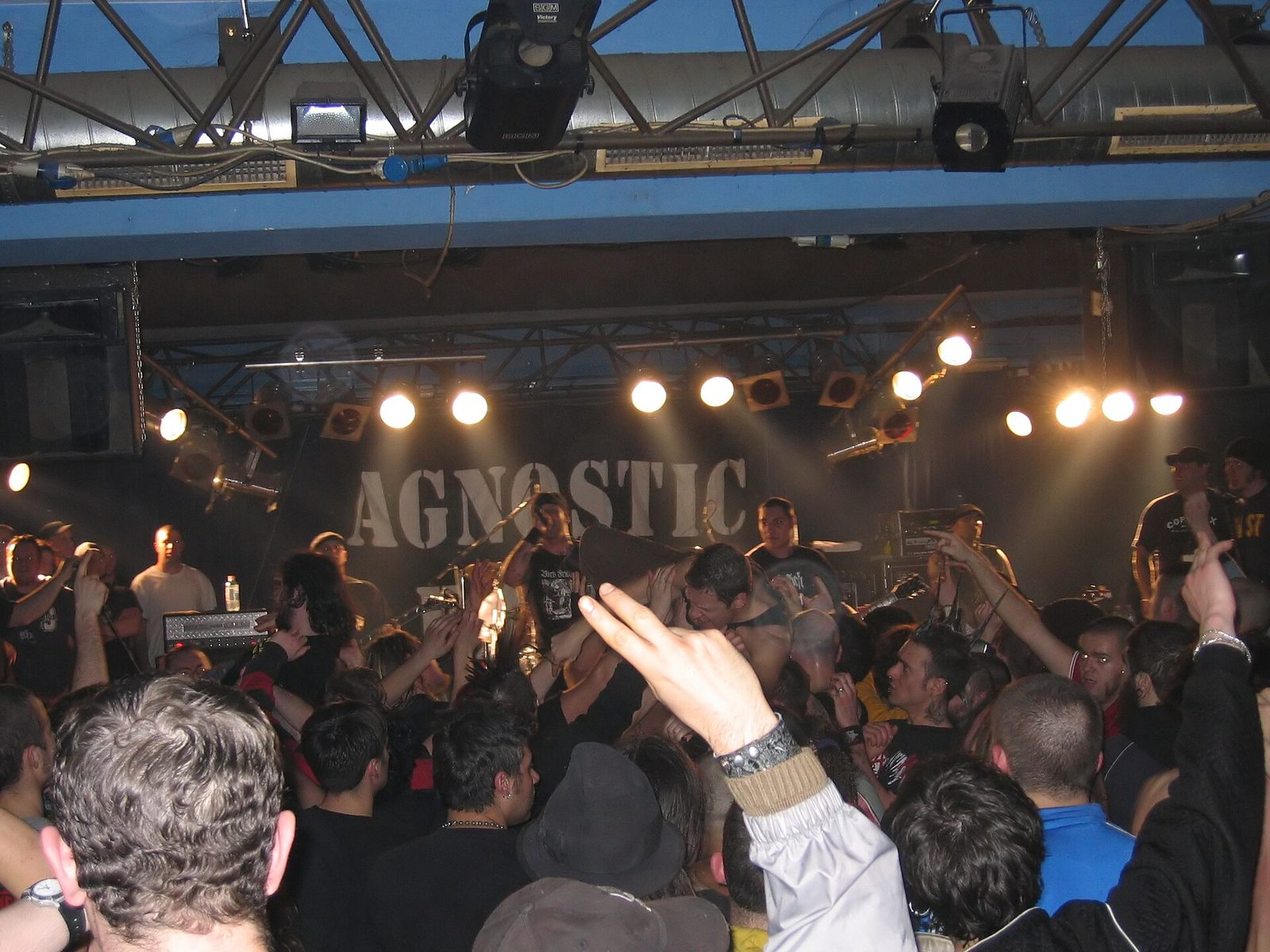 Agnostic Front on a club stage under a large stenciled AGNOSTIC banner while a shirtless fan crowd-surfs over a packed floor of raised hands