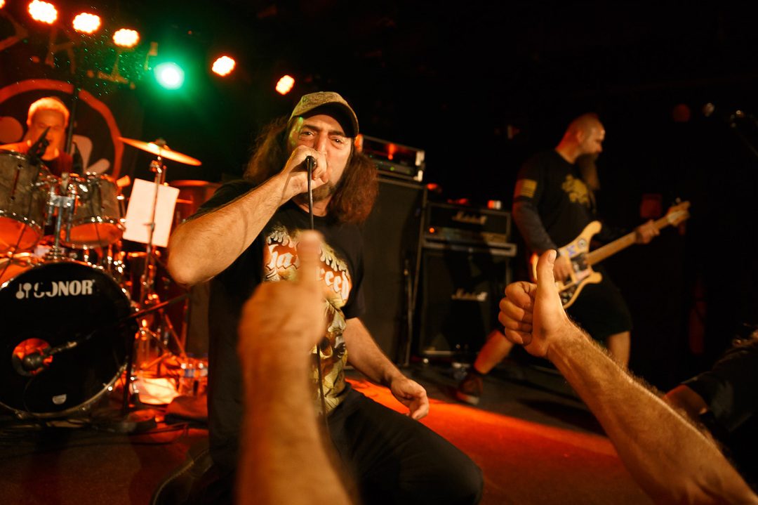 D.R.I.'s singer in a camouflage cap kneeling at the stage edge with the microphone as fans' thumbs-up hands reach toward him, the bassist and drummer behind