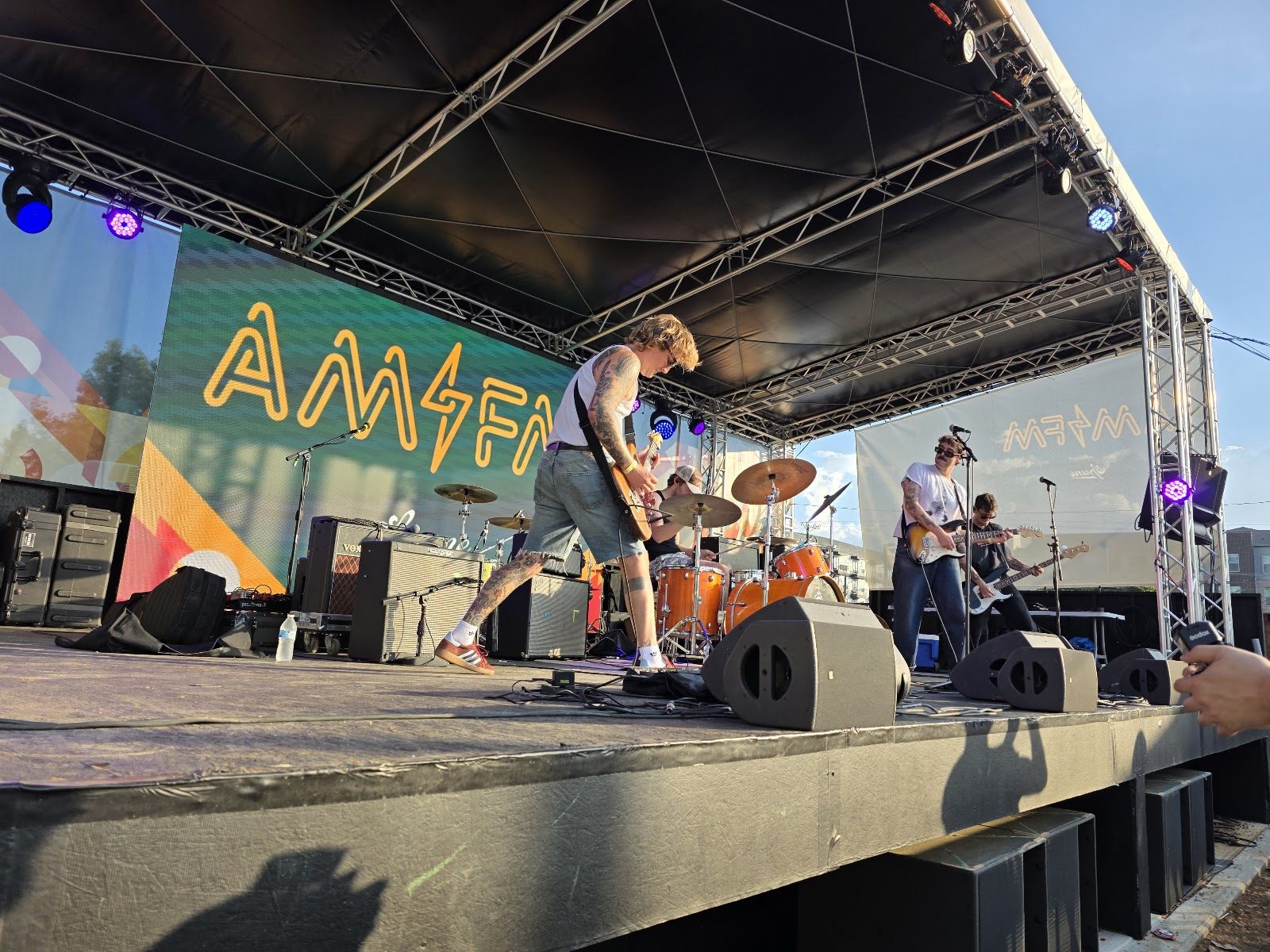 A rock band playing AM/FM's covered outdoor backyard stage in daylight, the AM/FM logo lit on a video wall behind them