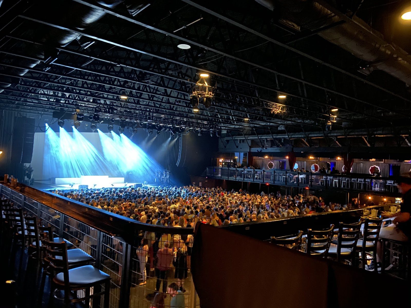 A packed general-admission floor at the Bomb Factory seen from the side balcony, blue stage lights beaming across a white backdrop under black steel trusses