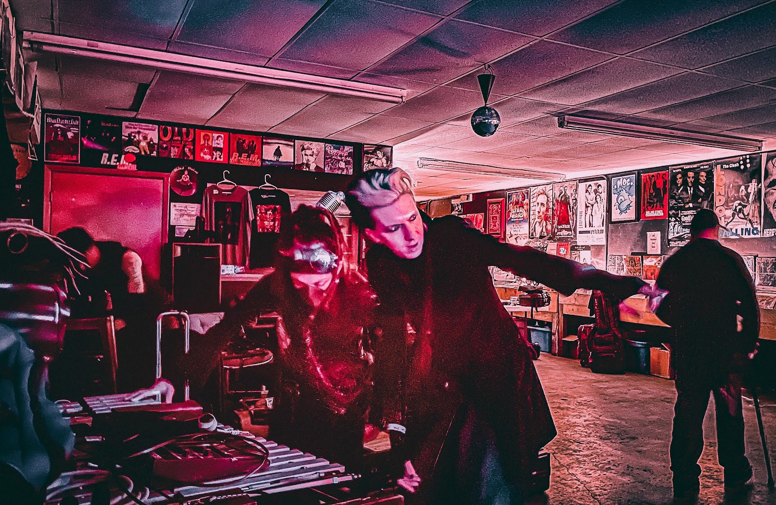 Two performers playing on the floor of Growl record store under red light, one with swept-back blond hair leaning forward mid-song, gig posters and record bins along the walls behind them