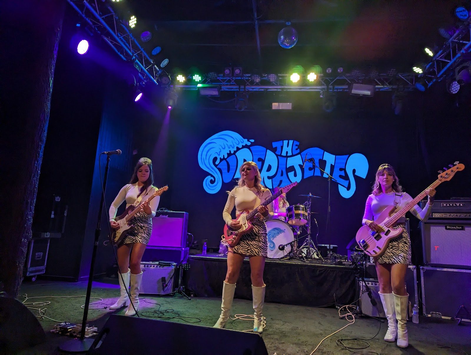 A three-piece band in white boots and zebra skirts playing guitars on Trees' stage under colored lights and a disco ball