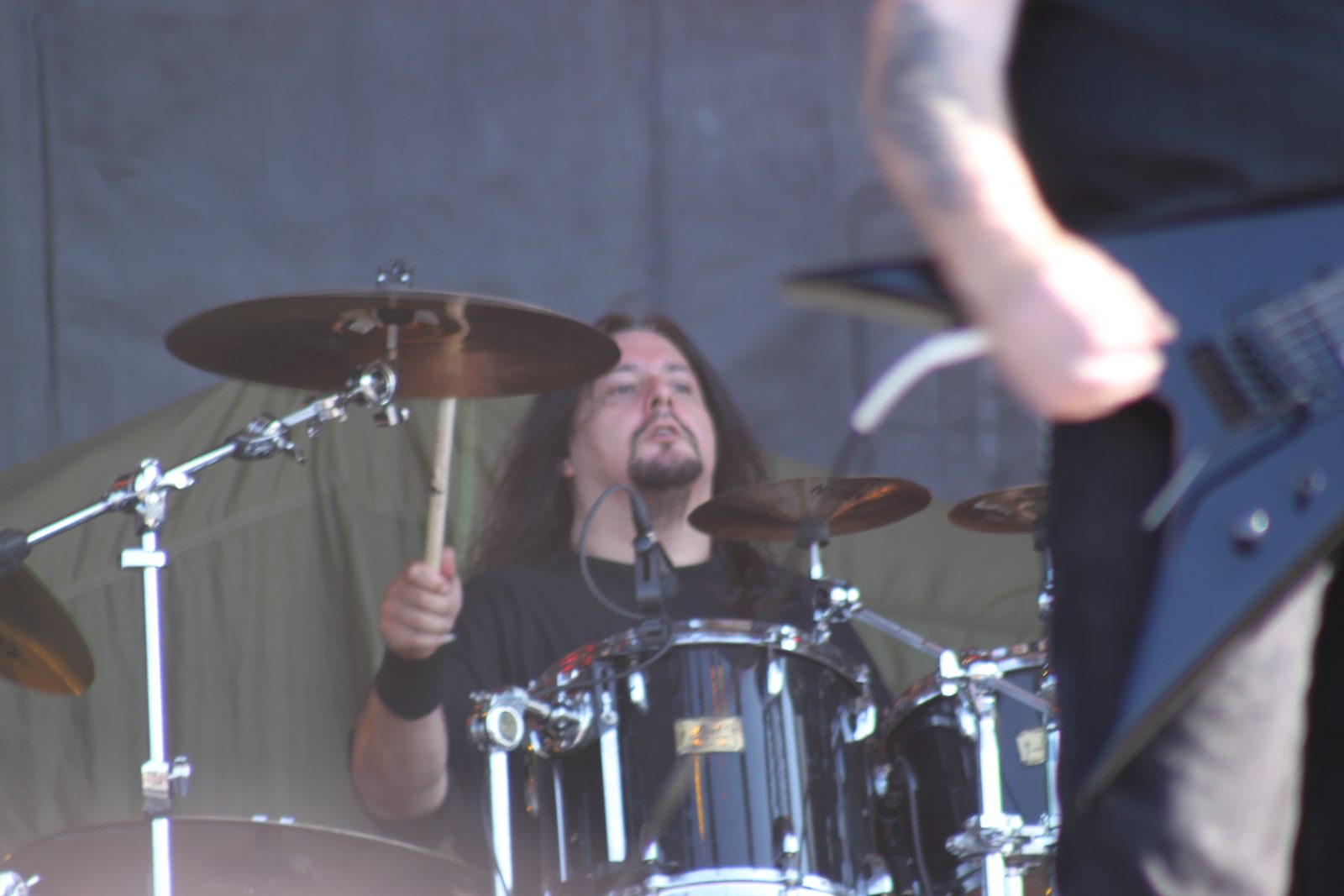 Gene Hoglan with long dark hair and a beard playing a drum kit on an outdoor stage, cymbals and a guitarist's arm in the foreground
