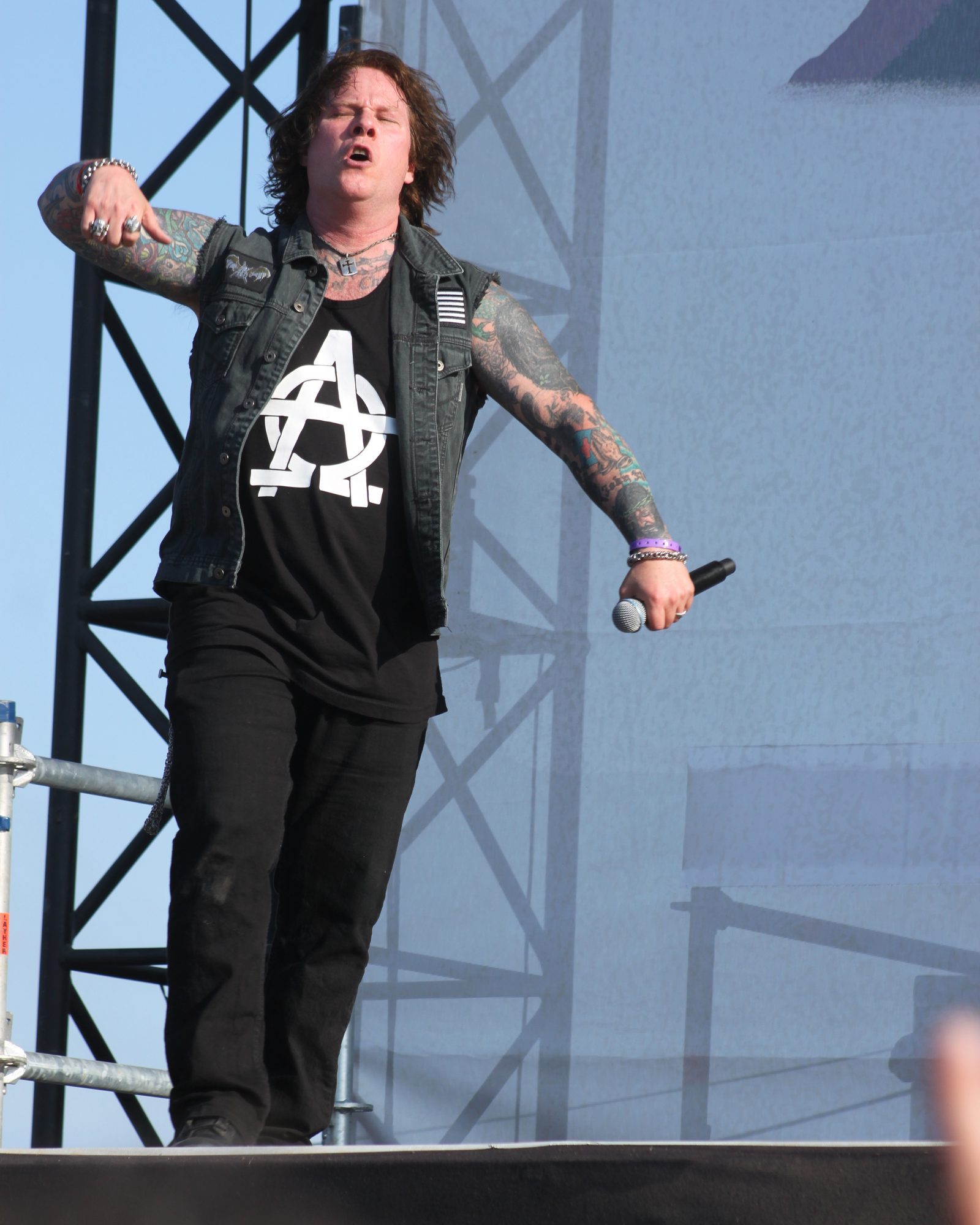 Ron Rinehart in a dark denim vest and anarchy-symbol T-shirt, tattooed arms outstretched, singing at the front of an outdoor festival stage