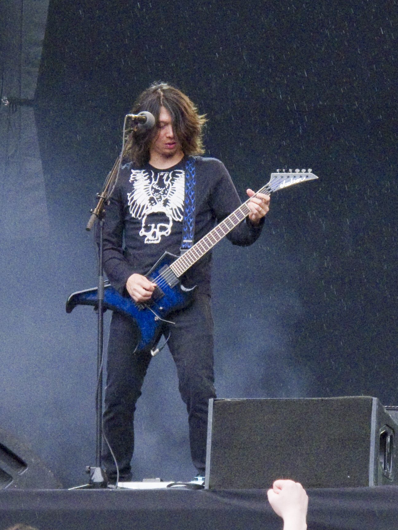 Rob Cavestany, with long dark hair over his face, playing a blue Jackson guitar in the rain on an outdoor stage in a black skull-print shirt