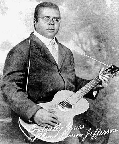 Black and white publicity portrait of Blind Lemon Jefferson in a suit, tie and small round glasses, seated with an acoustic guitar, signed 'Cordially Yours, Blind Lemon Jefferson'