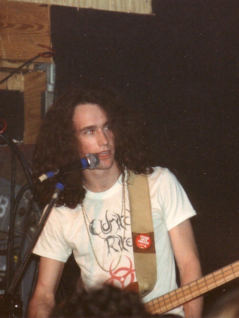 A young bassist with long dark curly hair and an Eldritch Rite T-shirt playing a Rickenbacker bass at a microphone
