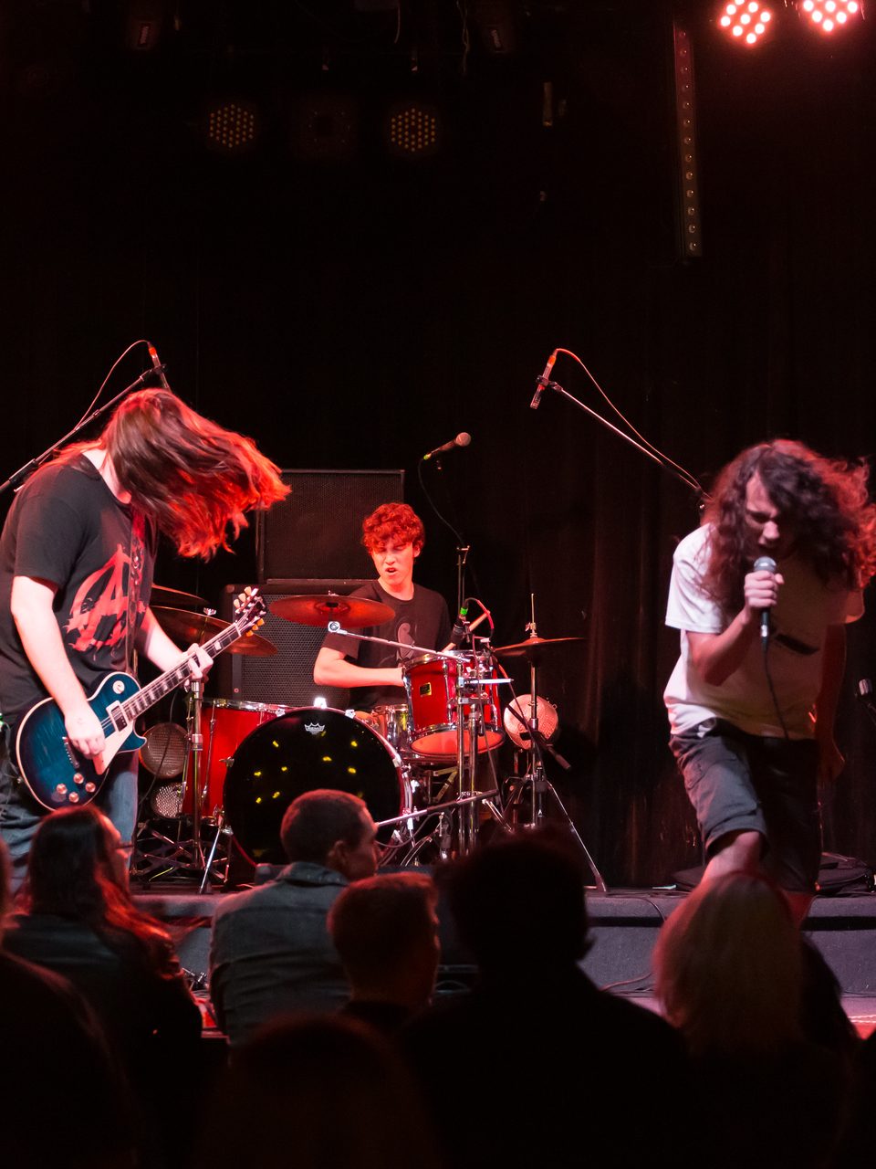 A young band playing under red lights on the Curtain Club stage, the guitarist's hair flying, the drummer behind him and the singer bent over his microphone, with the crowd in silhouette