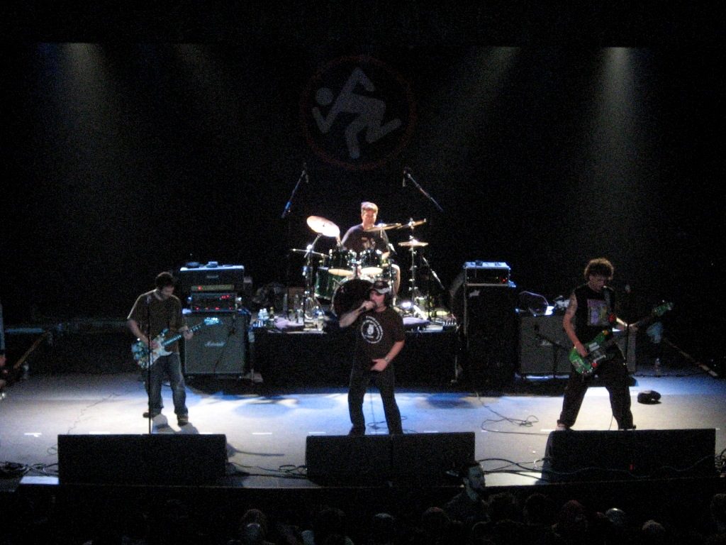 D.R.I. performing on a dark stage with the band's running-man logo on the backdrop, a singer at center and guitarists on each side