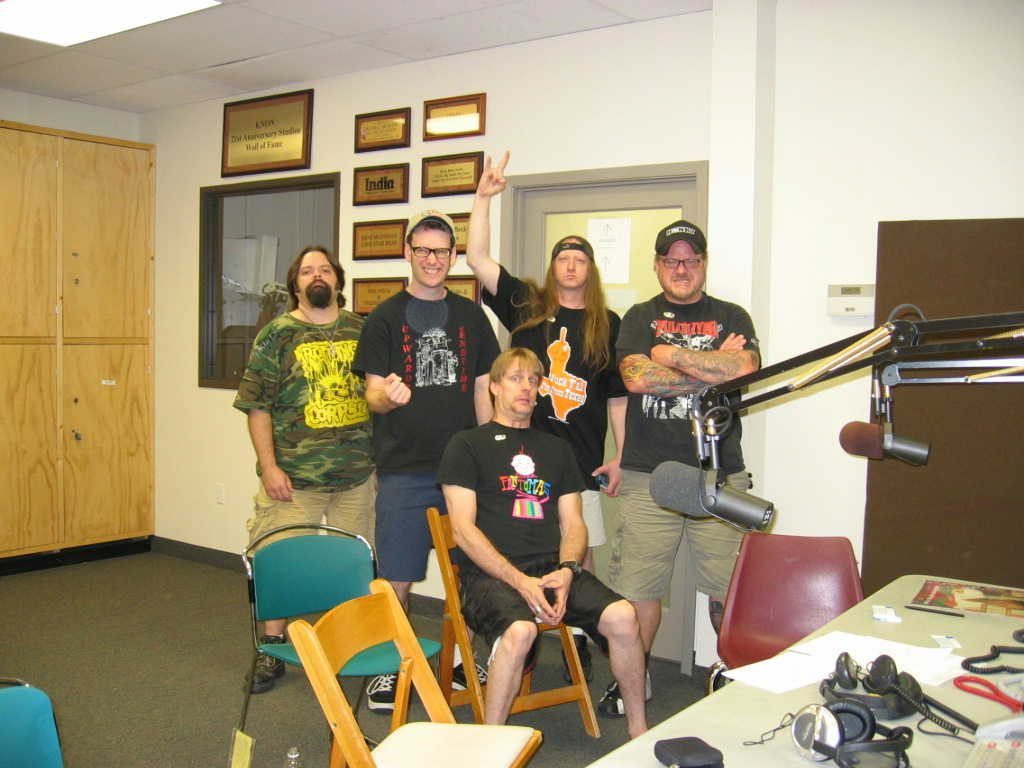 Five men in band T-shirts in a radio studio with framed plaques on the wall and a microphone on a boom arm in the foreground, one seated in front