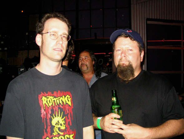 Two men at a dark club, one in glasses and a Rotting Corpse T-shirt, the other in a cap holding a beer bottle