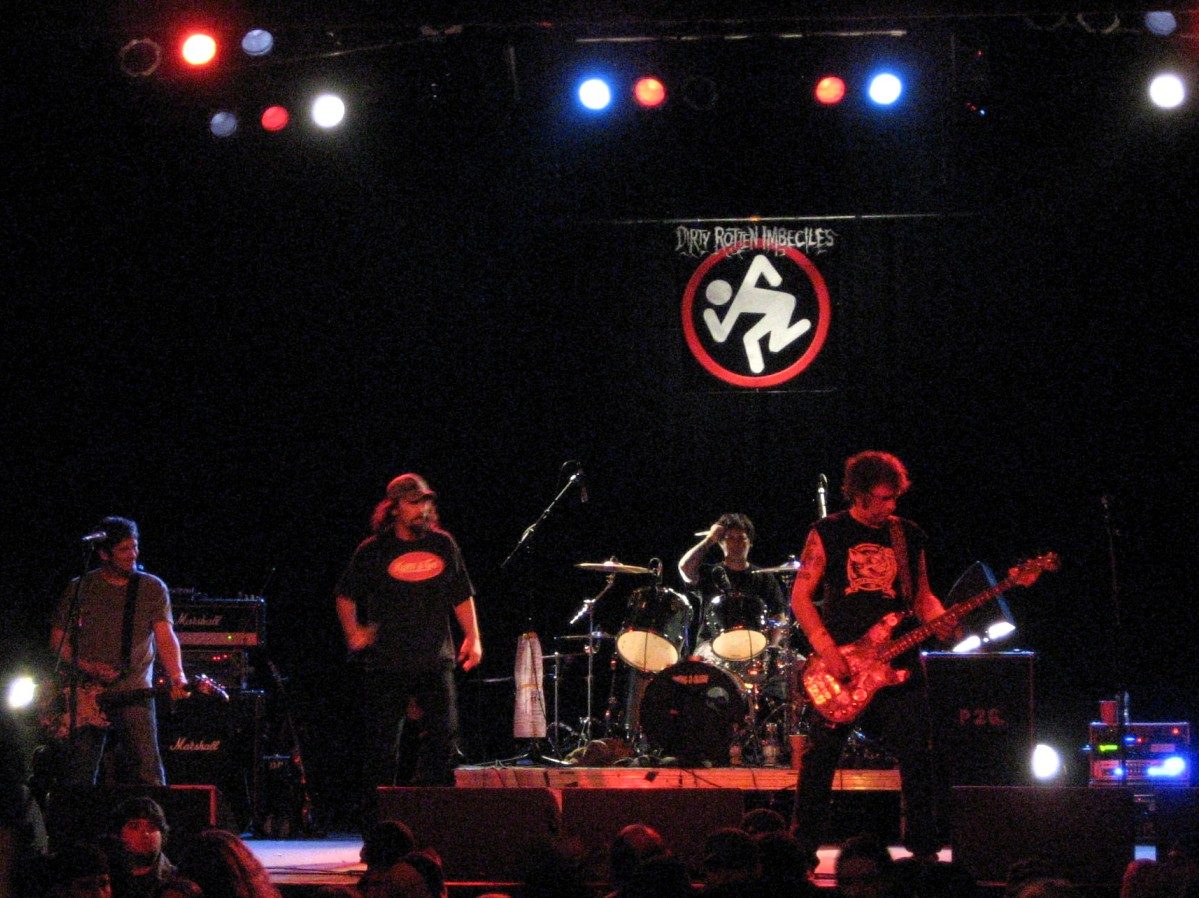 D.R.I. on a dark stage with the bassist, the singer in a cap, the drummer and the guitarist, under a black banner with the band's white running stick figure inside a red ring