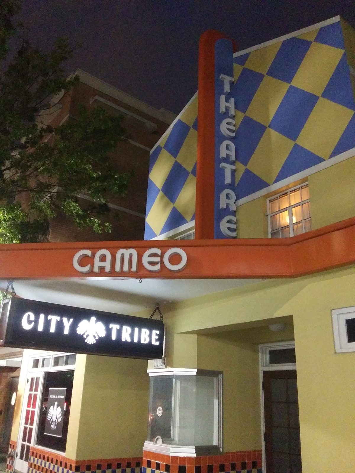 The Cameo Theatre building at night: an orange CAMEO marquee, a vertical THEATRE sign on a blue-and-yellow diamond facade, and a lit City Tribe sign hanging below