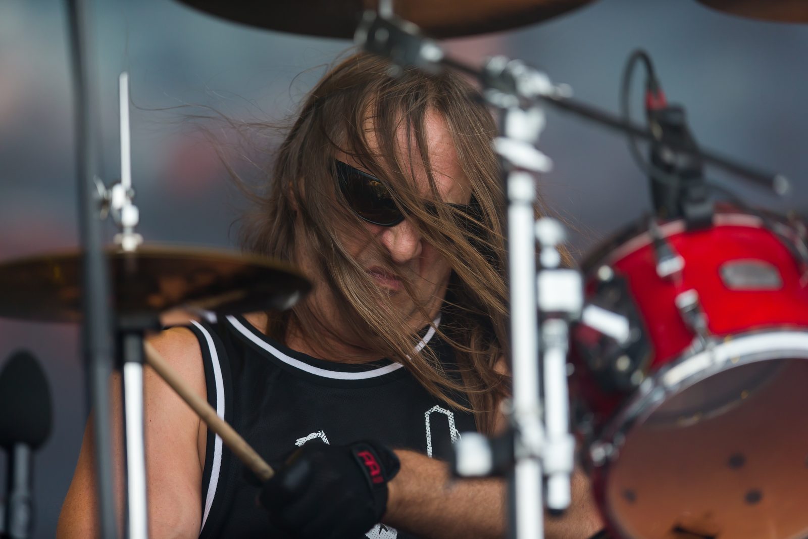 Tom Hunting in dark sunglasses with long hair swinging across his face, playing a red drum kit on an outdoor stage