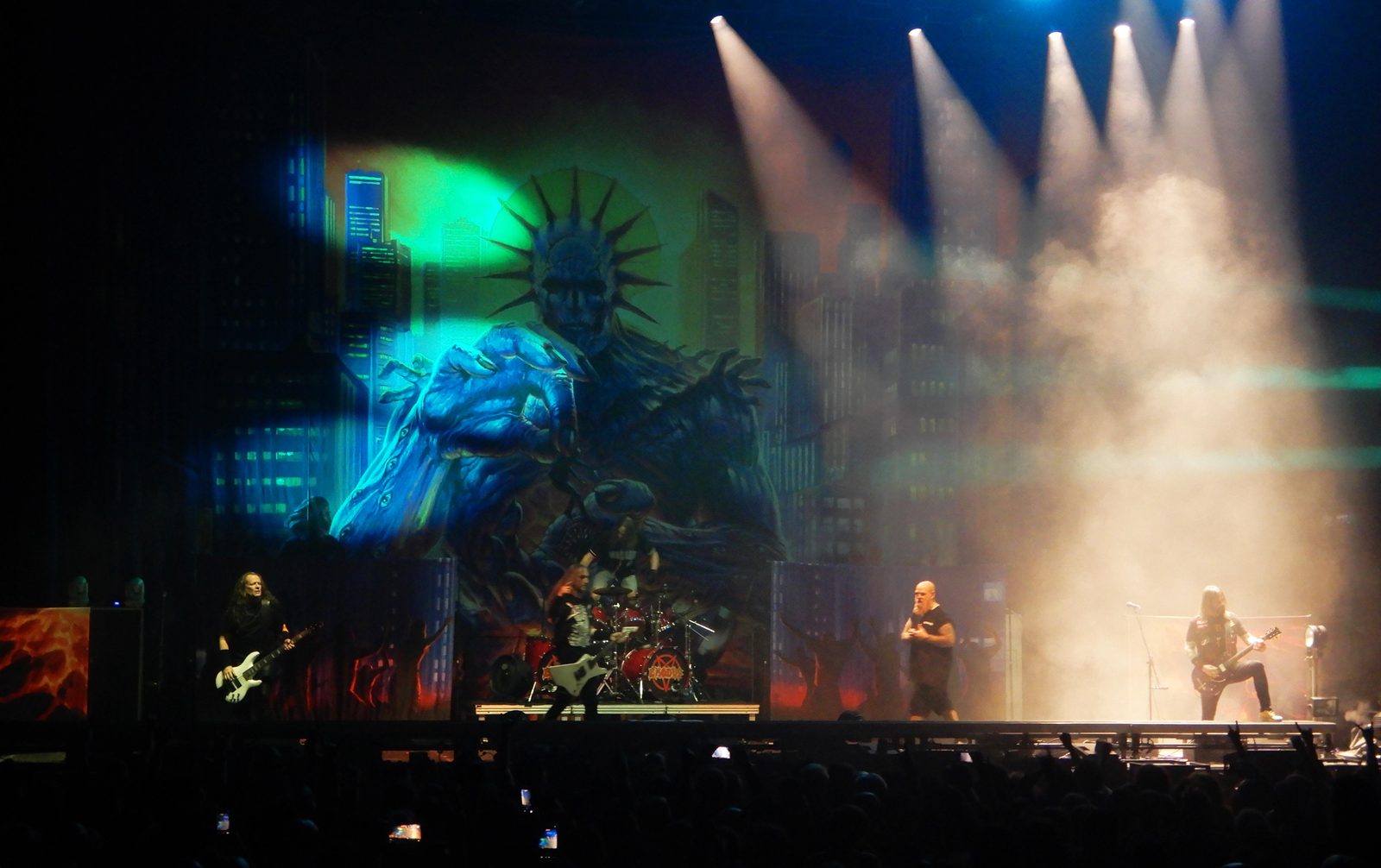 Exodus on a wide arena stage in Paris, a bassist at left, a bald singer and a guitarist at right in stage smoke, a giant painted Statue of Liberty figure on the backdrop
