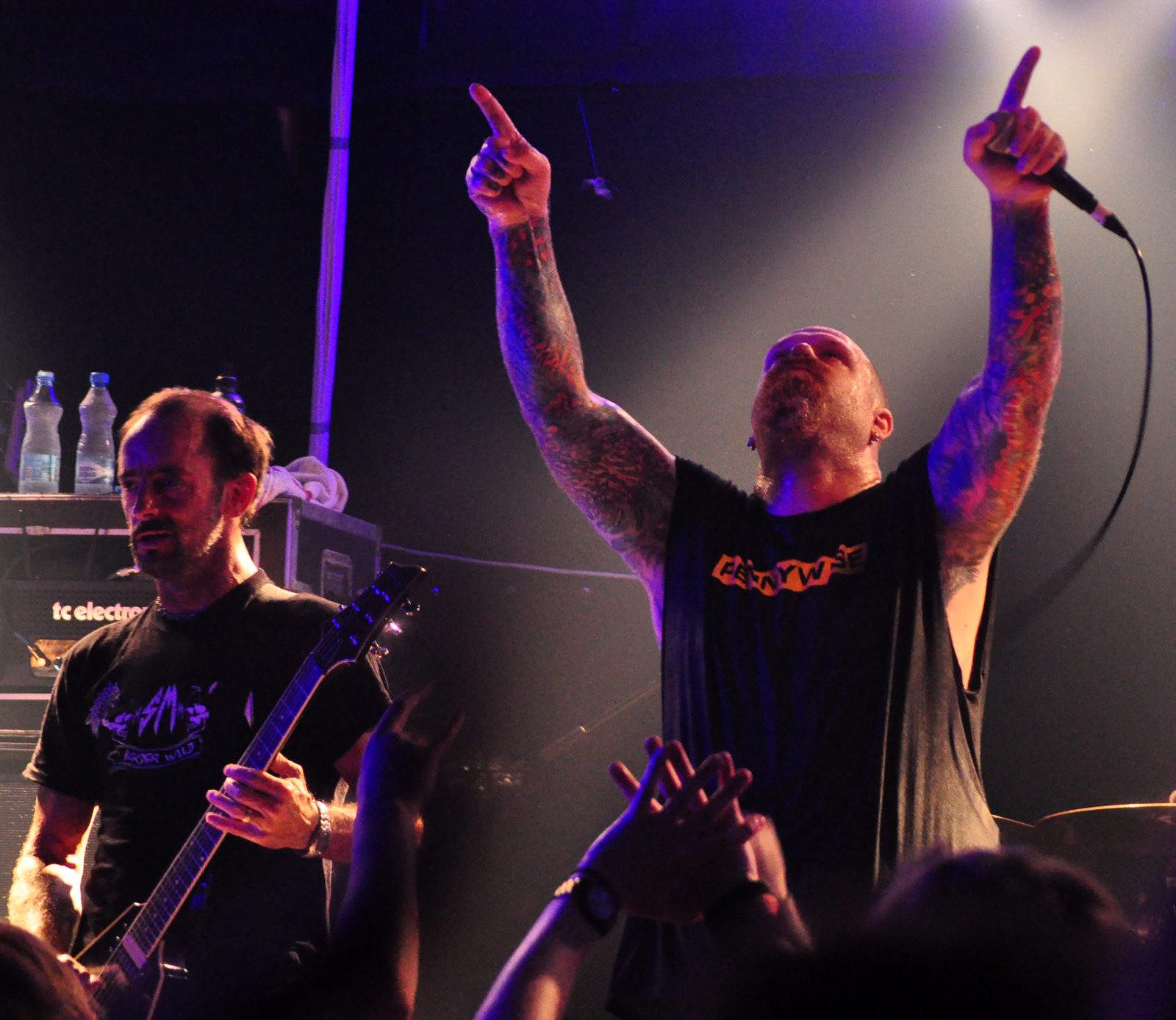 Rick Hunolt playing a black guitar at left while a bald, tattooed singer raises both arms and points upward on a dark club stage, fans' hands raised in front