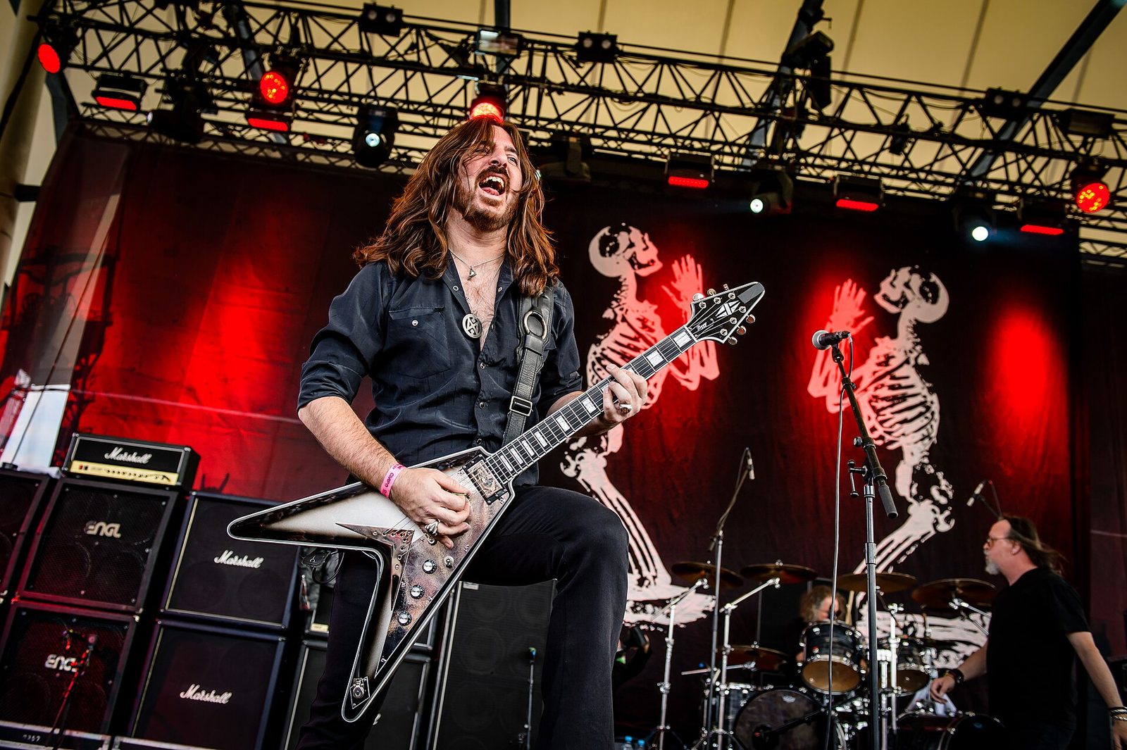 Cirith Ungol's long-haired guitarist plays a flying-V at the front of a festival stage under red lights, with skeleton banners and the drummer behind him