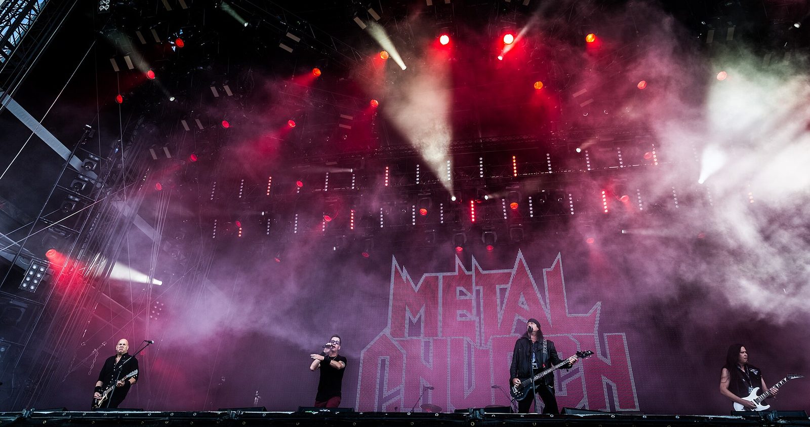 Metal Church plays a huge outdoor festival stage, four musicians small in front of a giant red Metal Church logo under smoke and red lights
