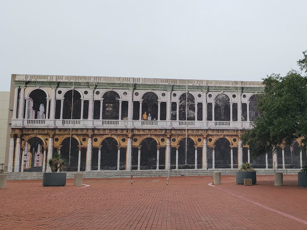 A long two-storey building at the Dallas Communications Complex in Irving, its wall painted as a Renaissance arcade of white columns and arches, behind an empty red-brick plaza with planters and a large oak tree