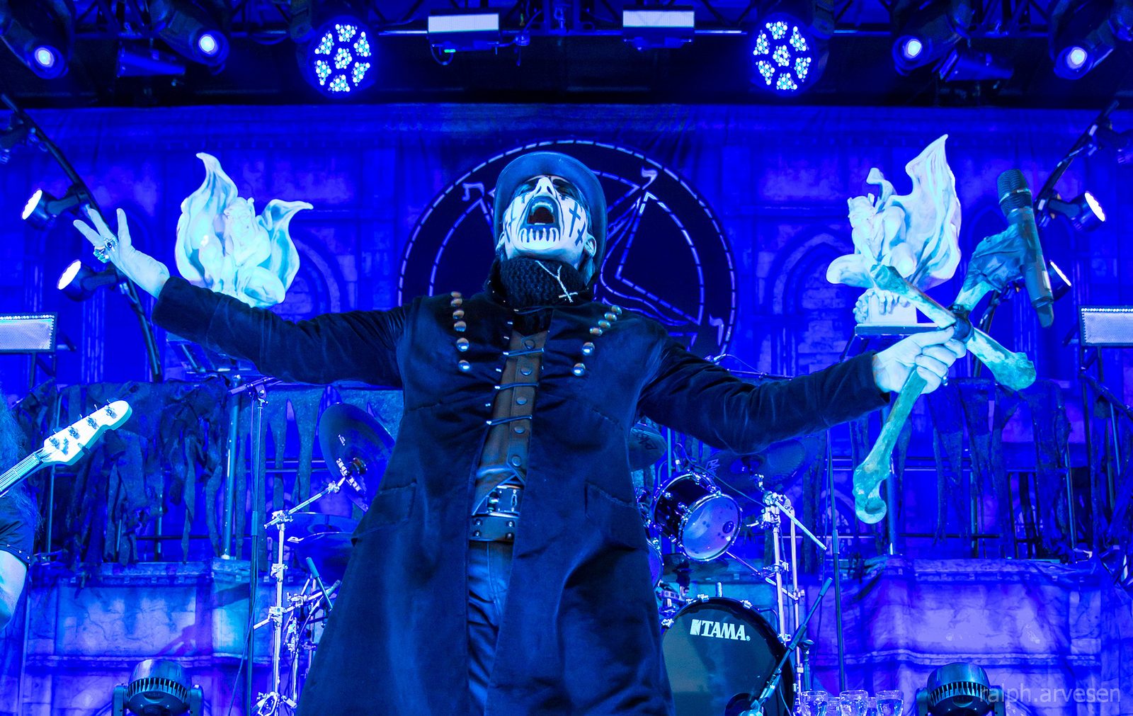 King Diamond in black-and-white face paint and a long dark coat, arms spread wide on a blue-lit stage with a drum kit and pale flame props behind him