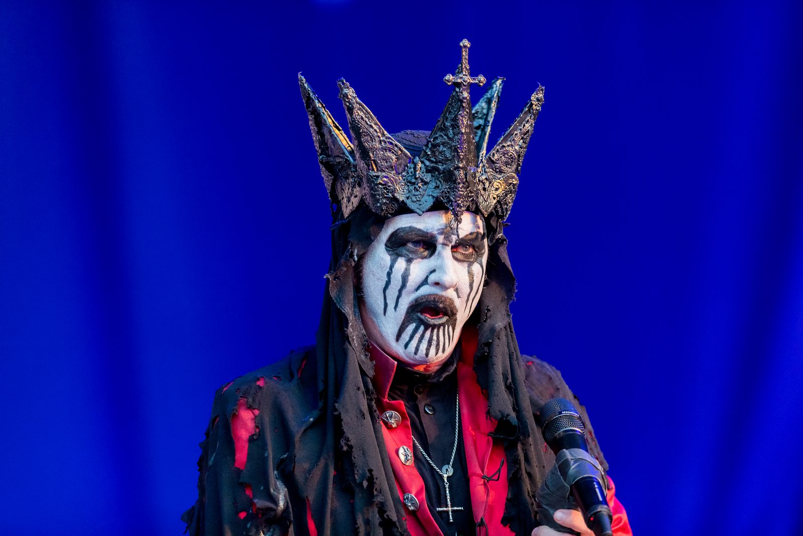 King Diamond in white face paint with black markings and a tall spiked silver headpiece, singing into a microphone against a deep blue backdrop