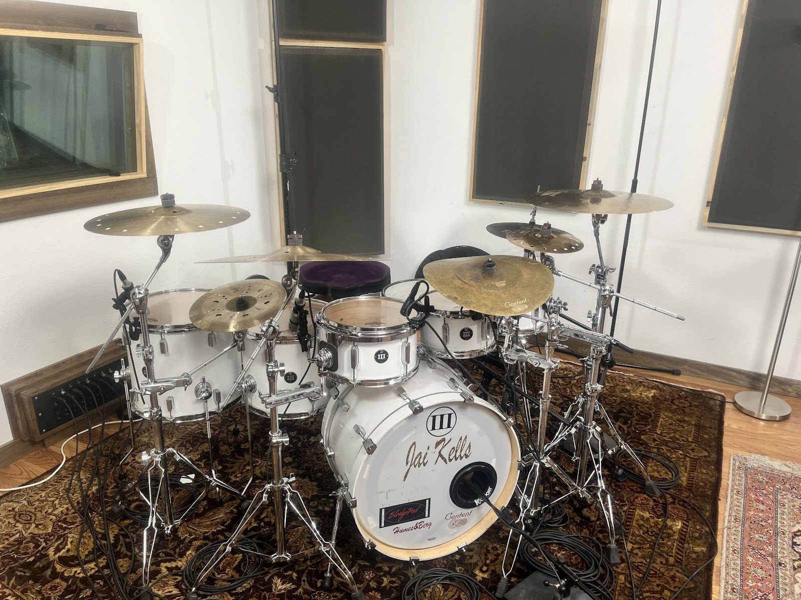 A white drum kit with brass cymbals and close microphones set up on a patterned rug in a studio live room, with dark acoustic panels on white walls and a control-room window at left