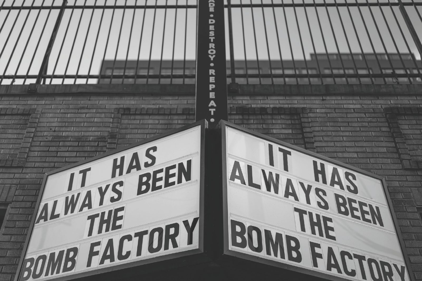Black and white photo of a double-sided marquee on a brick wall reading It Has Always Been The Bomb Factory, under a tall iron fence