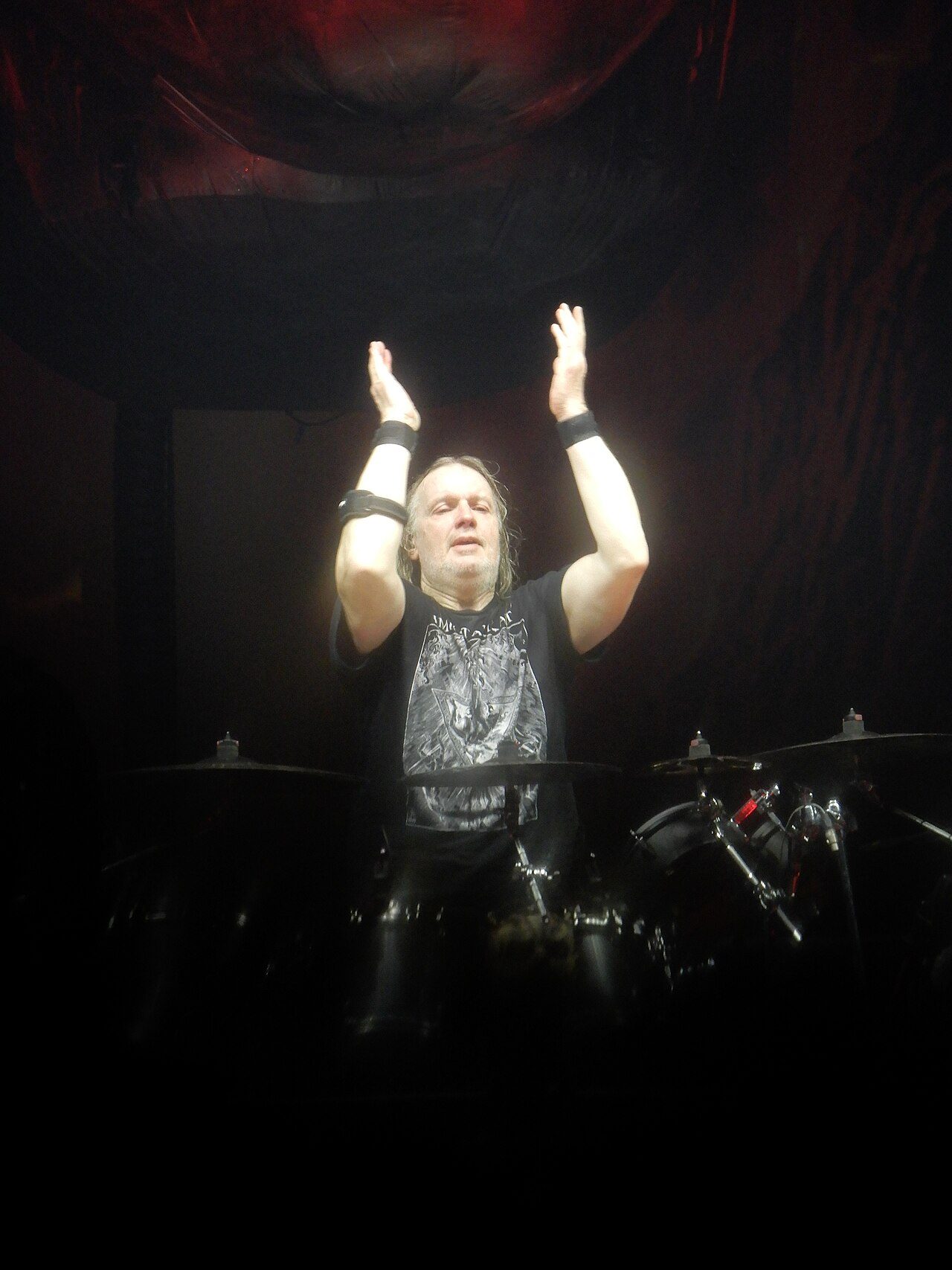 Ventor, gray-haired and bearded in a black band T-shirt, raising both hands above his head behind his drum kit under a single spotlight