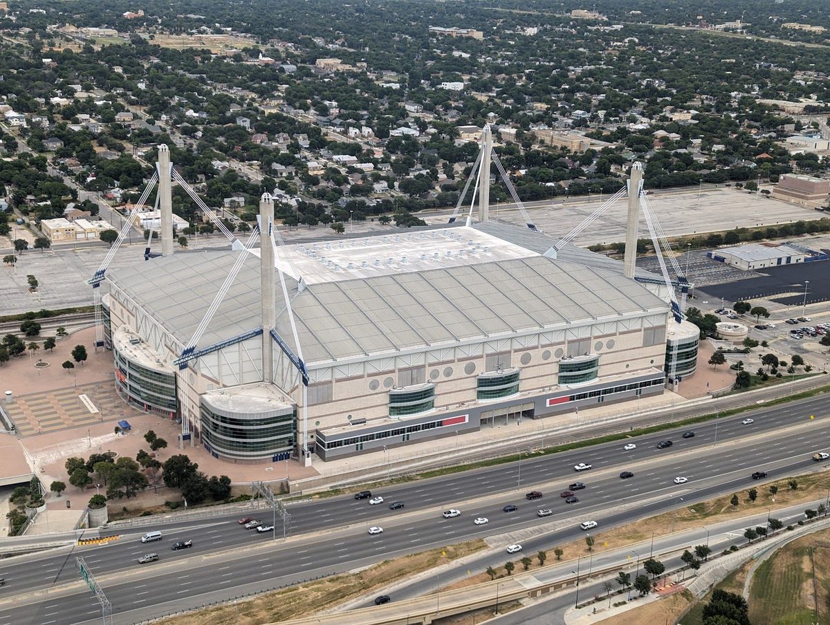 Aerial view of the Alamodome in San Antonio, a white-roofed stadium with four tall corner masts and support cables, beside a busy freeway