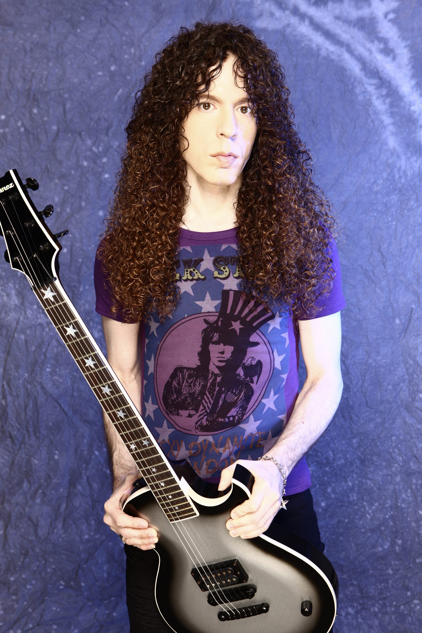 Marty Friedman with long curly dark hair, wearing a purple T-shirt and holding a silver and black guitar against a blue backdrop
