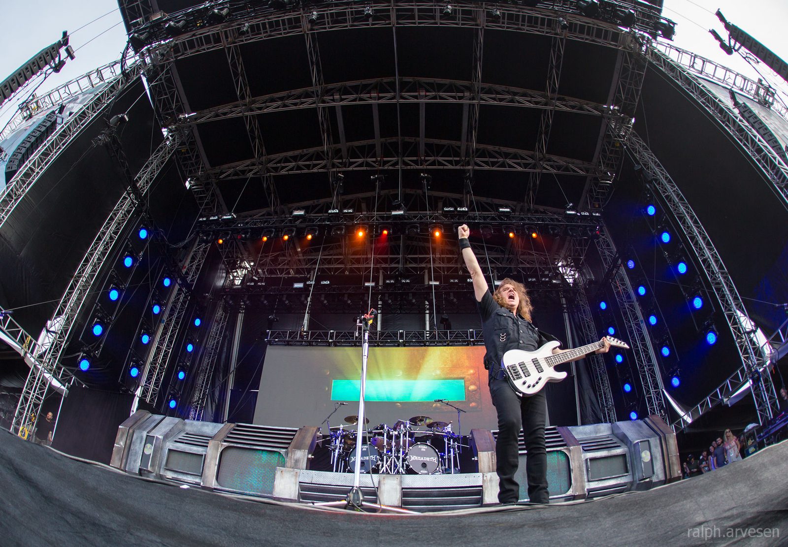 Wide-angle view of a large outdoor stage under a steel roof, a bassist with a white bass raising his fist at the front edge, drums and blue lights behind him