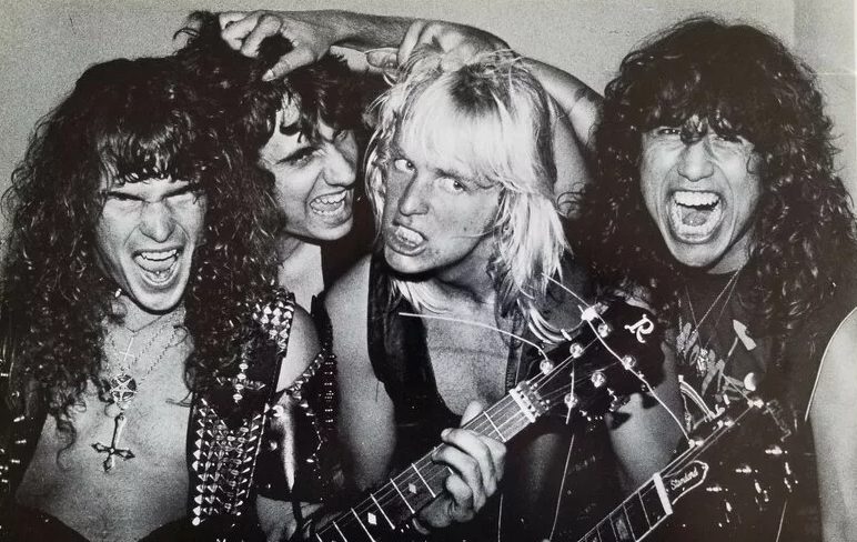 Black-and-white 1983 photo of Slayer's four members crowded together and snarling at the camera, one holding a guitar, two wearing studded leather