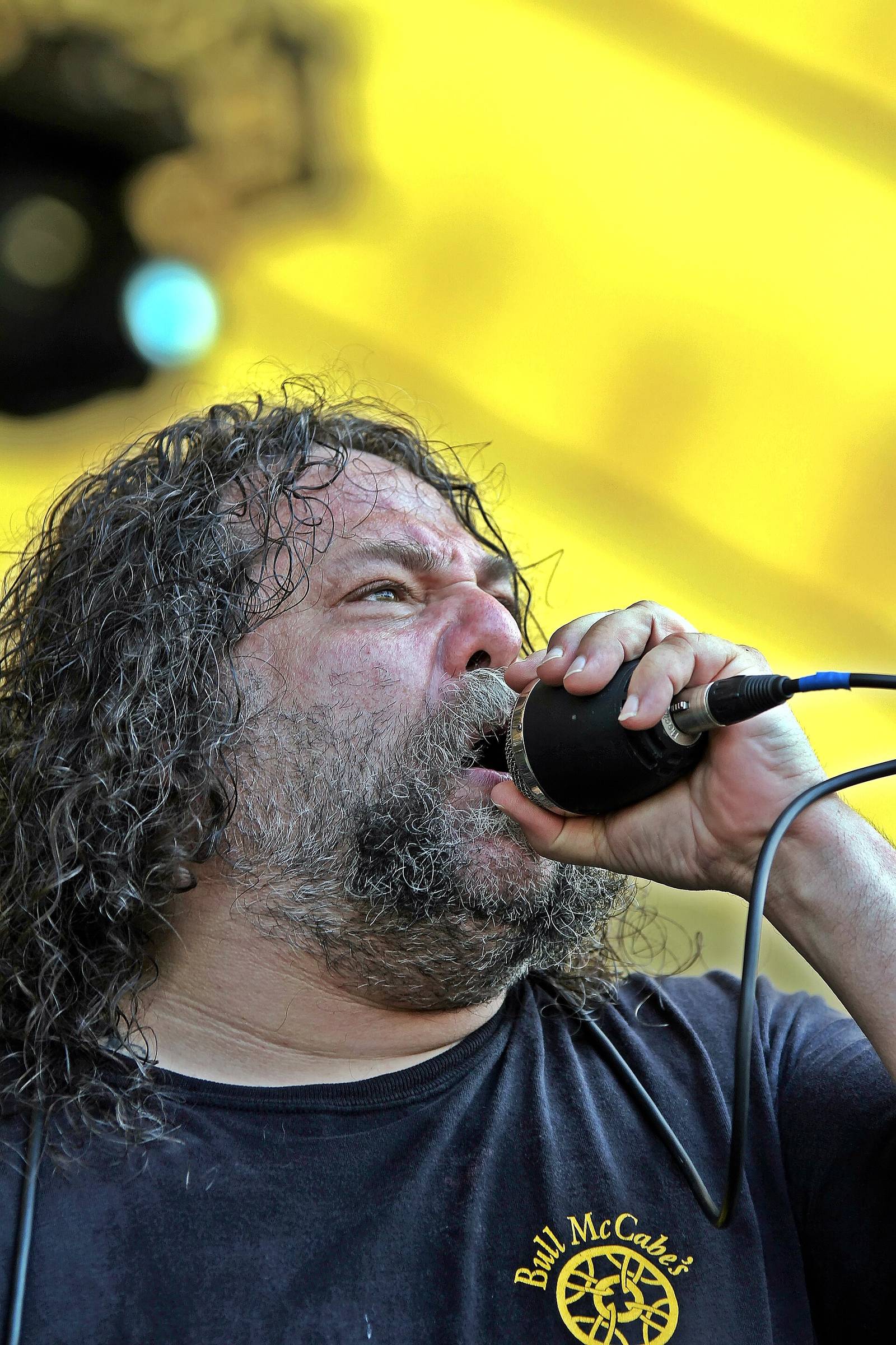 Close-up of Billy Milano singing into a handheld microphone, head tilted up, with long curly gray-streaked hair and a beard, against a bright yellow stage backdrop