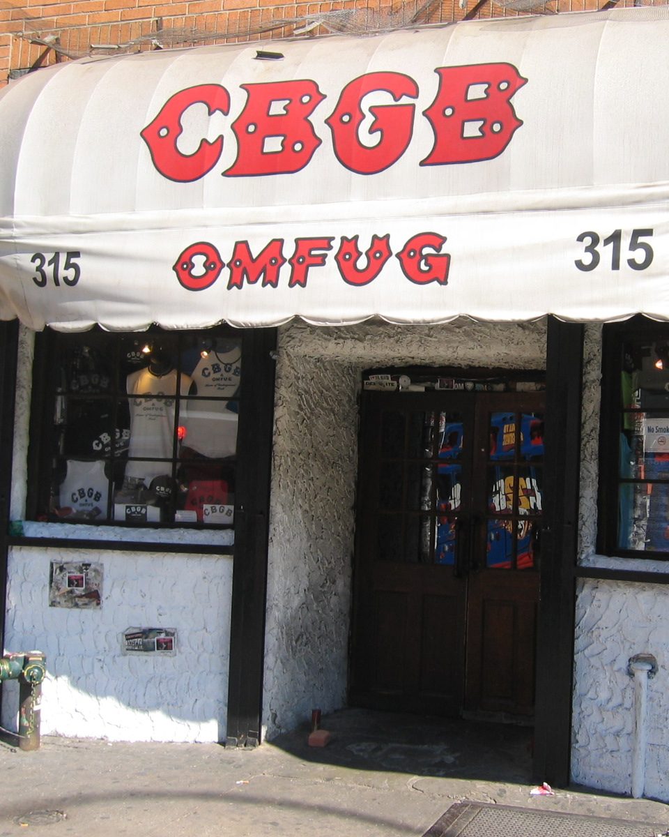 The front of CBGB on the Bowery: a white rounded awning reading CBGB OMFUG in red letters with the number 315 on each side, over a rough white wall, two windows of T-shirts and a dark wooden double door