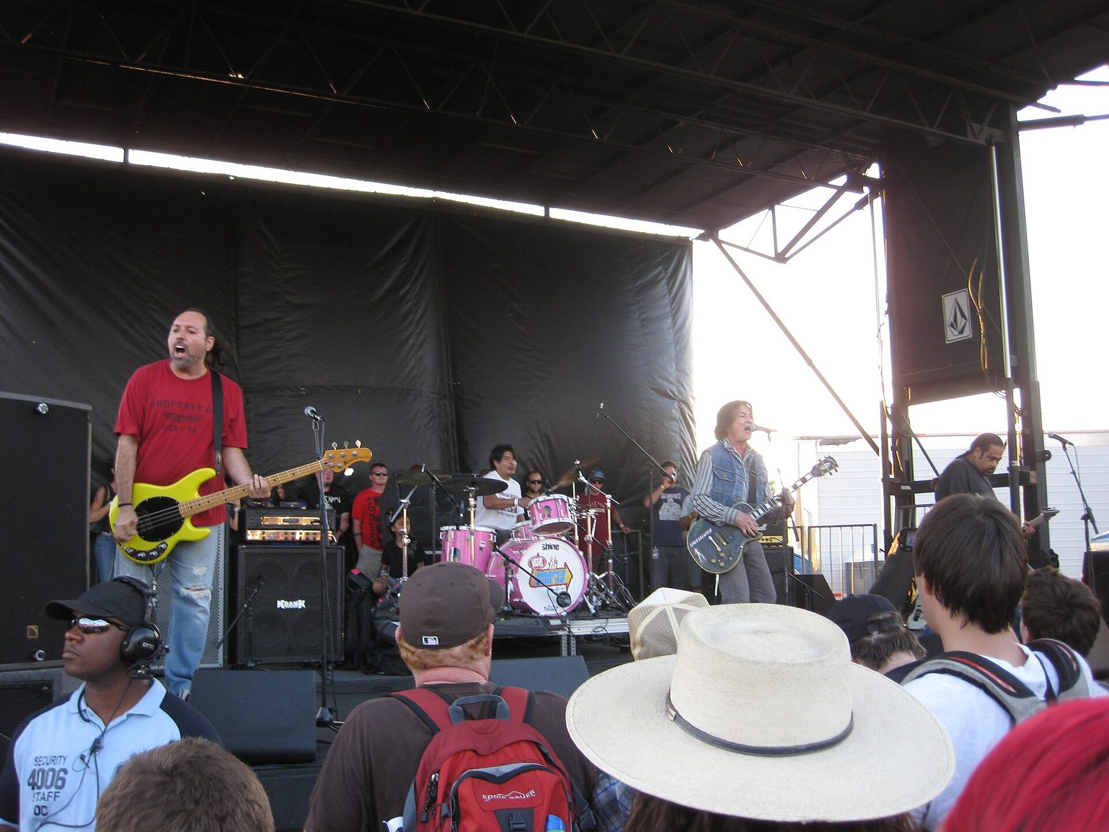 The Los Angeles punk band Fear playing an outdoor stage, a bassist in a red shirt with a yellow bass on the left and Lee Ving singing at a microphone with a guitar on the right, fans in hats in the foreground