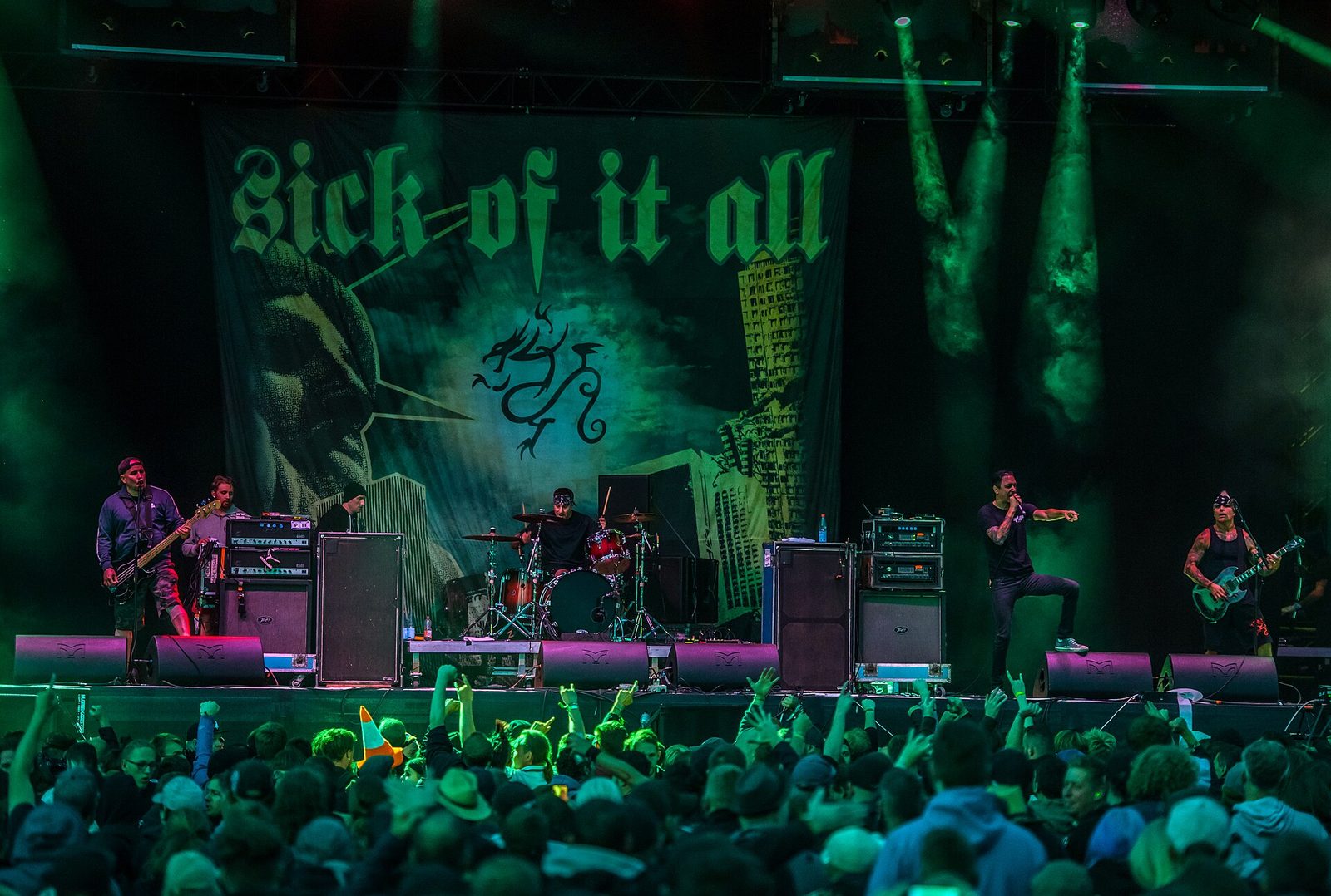 Sick of It All on a festival stage in front of a large green backdrop with the band's name in blackletter, the singer pointing at the crowd, and fans raising their hands in front of the stage