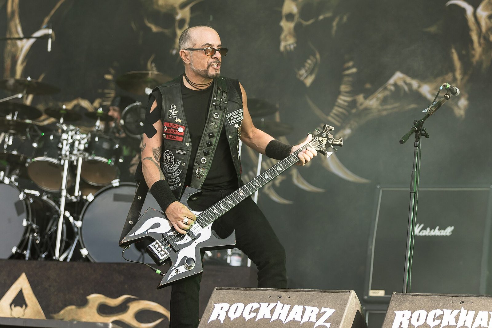 D.D. Verni in sunglasses and a patched black leather vest playing a black and white star-shaped bass in front of a Marshall cabinet, a skull backdrop behind him